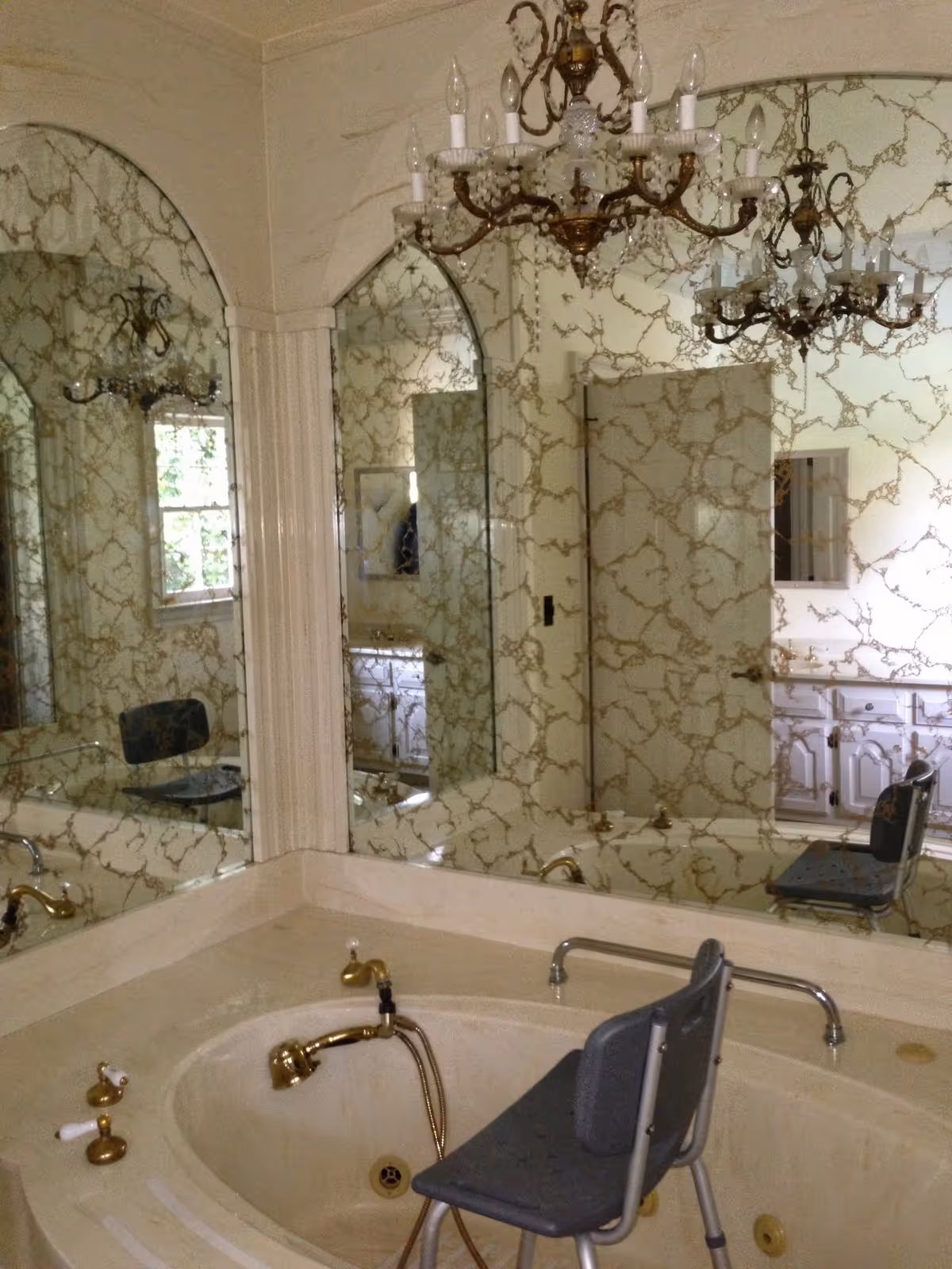 A bathroom with a large corner bathtub featuring gold-colored fixtures and a gray shower chair inside. The walls are covered with decorative mirrors that have a marbled pattern, reflecting a chandelier hanging from the ceiling and a window. A vanity with a sink and a mirror is visible in the background.