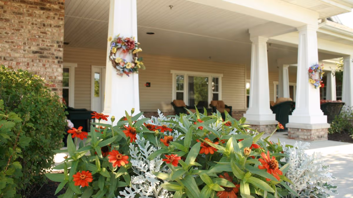 Outdoor view of a senior living facility porch area with white columns decorated with floral wreaths. There are red flowers and green plants in the foreground, and cushioned chairs are visible on the porch under a covered roof.