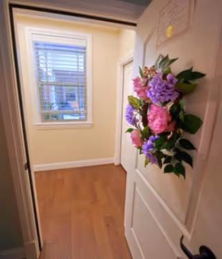 View through an open door into a small room with wooden flooring and a window with white blinds. A colorful floral wreath with pink and purple flowers is hanging on the door.