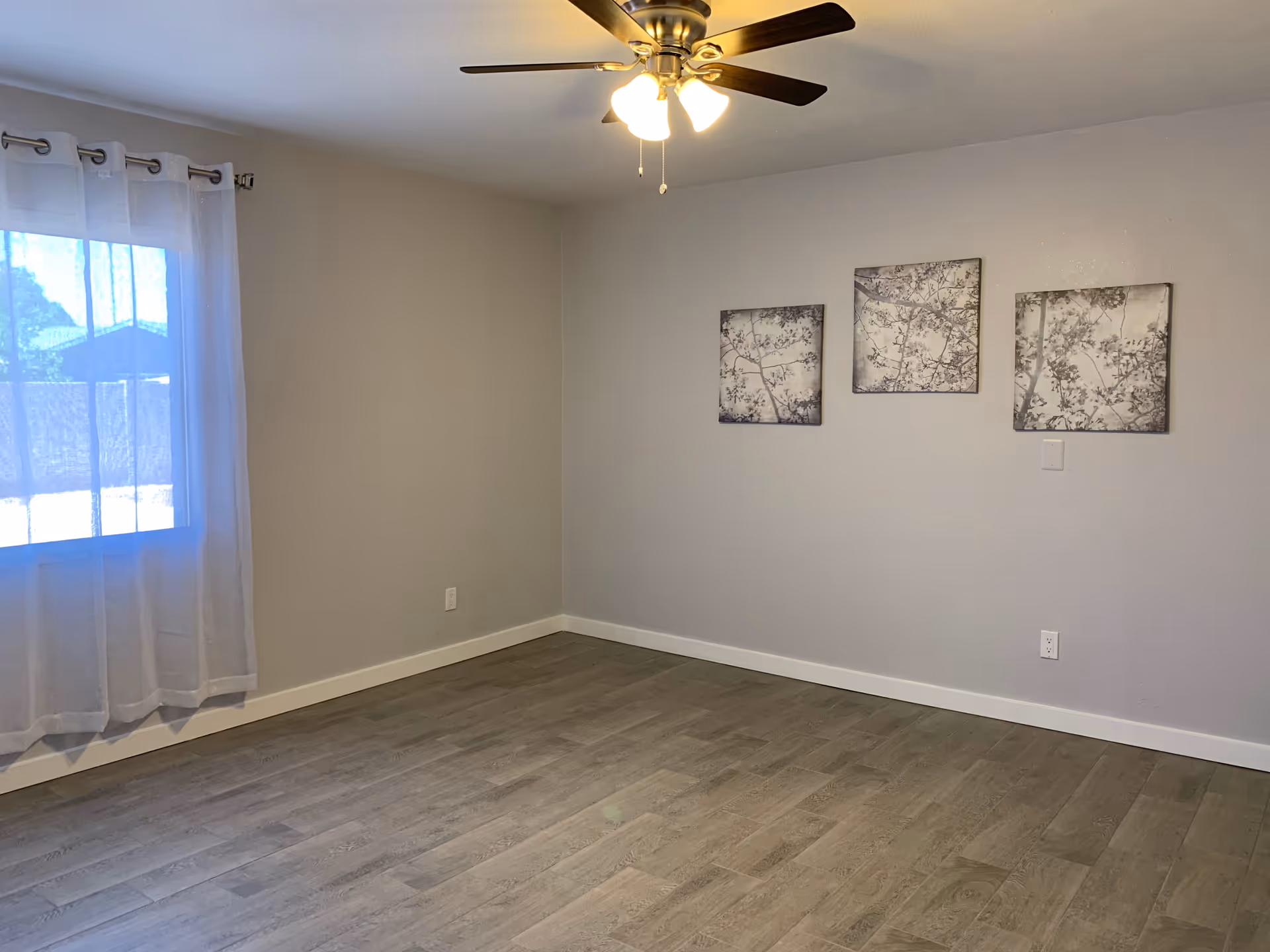 Empty room with light gray walls, wood-look flooring, a ceiling fan with lights, a window with sheer white curtains, and three framed pictures of tree branches on the wall.
