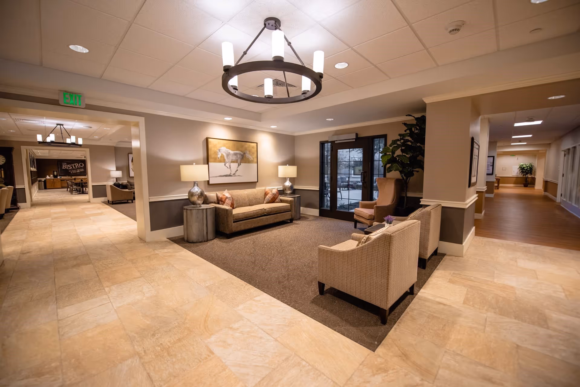 A cozy seating area in a senior living facility with a sofa, two armchairs, side tables with lamps, and a large painting of a horse on the wall. The area is carpeted and adjacent to a hallway with tile flooring. There is a glass door leading outside and a chandelier hanging from the ceiling. In the background, a dining area labeled 'Bistro' is visible.