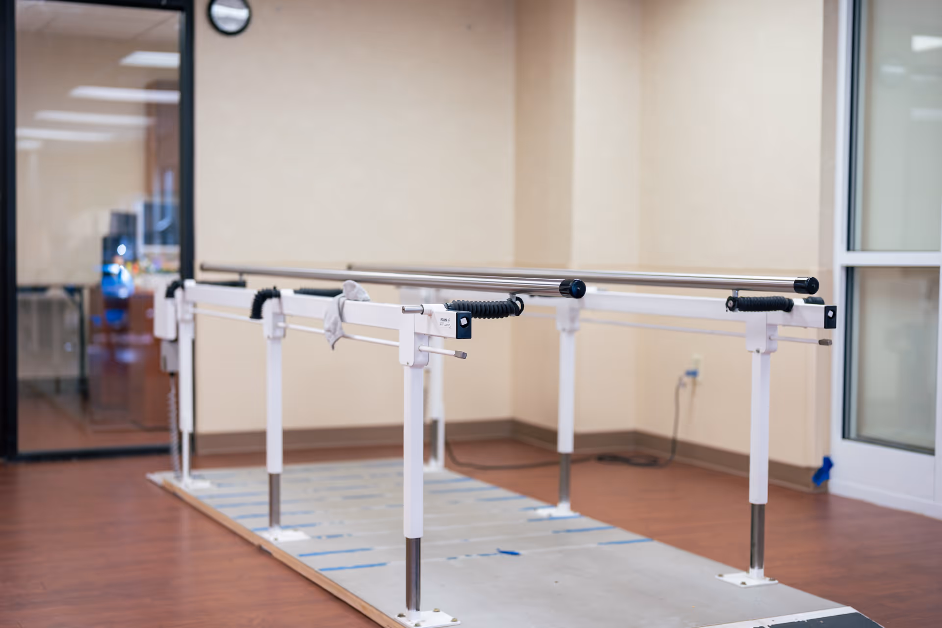 A rehabilitation room featuring parallel bars used for physical therapy and walking exercises, with a clock on the wall and a glass door in the background.
