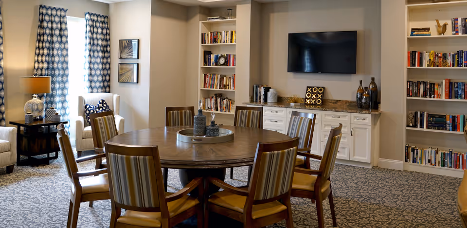 A cozy living room area with a round wooden table surrounded by six striped cushioned chairs. The room features built-in white bookshelves filled with books and decorative items, a flat-screen TV mounted on the wall above a white cabinet with a granite countertop, and two armchairs near a window with patterned blue curtains. The floor is carpeted with a patterned design, and a table lamp sits on a small side table next to one of the armchairs.