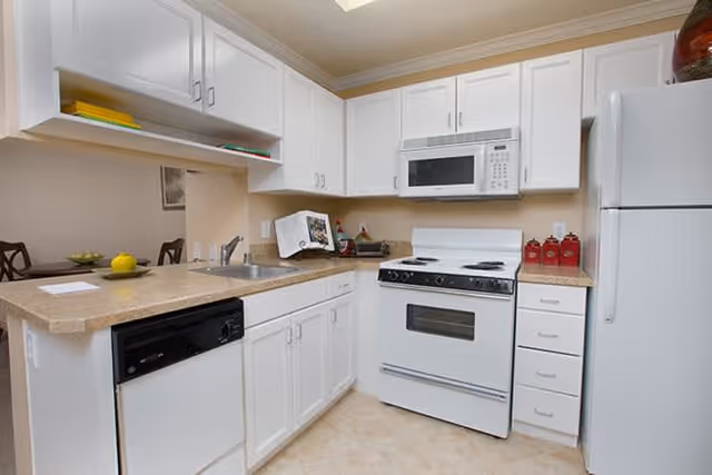 A clean and well-lit kitchen with white cabinets, a white refrigerator, a white microwave mounted above a white stove, a dishwasher, and a countertop with a sink. The kitchen opens into a dining area with a table and chairs visible in the background.