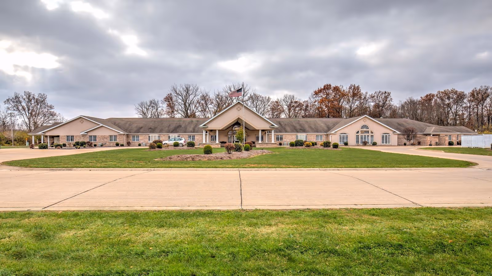 Wide front exterior view of Brookstone Estates of Paris, a single-story senior living facility with a large driveway and well-maintained lawn. The building has a beige facade with multiple windows and an American flag flying on a flagpole in front. Trees with autumn foliage are visible in the background under a cloudy sky.
