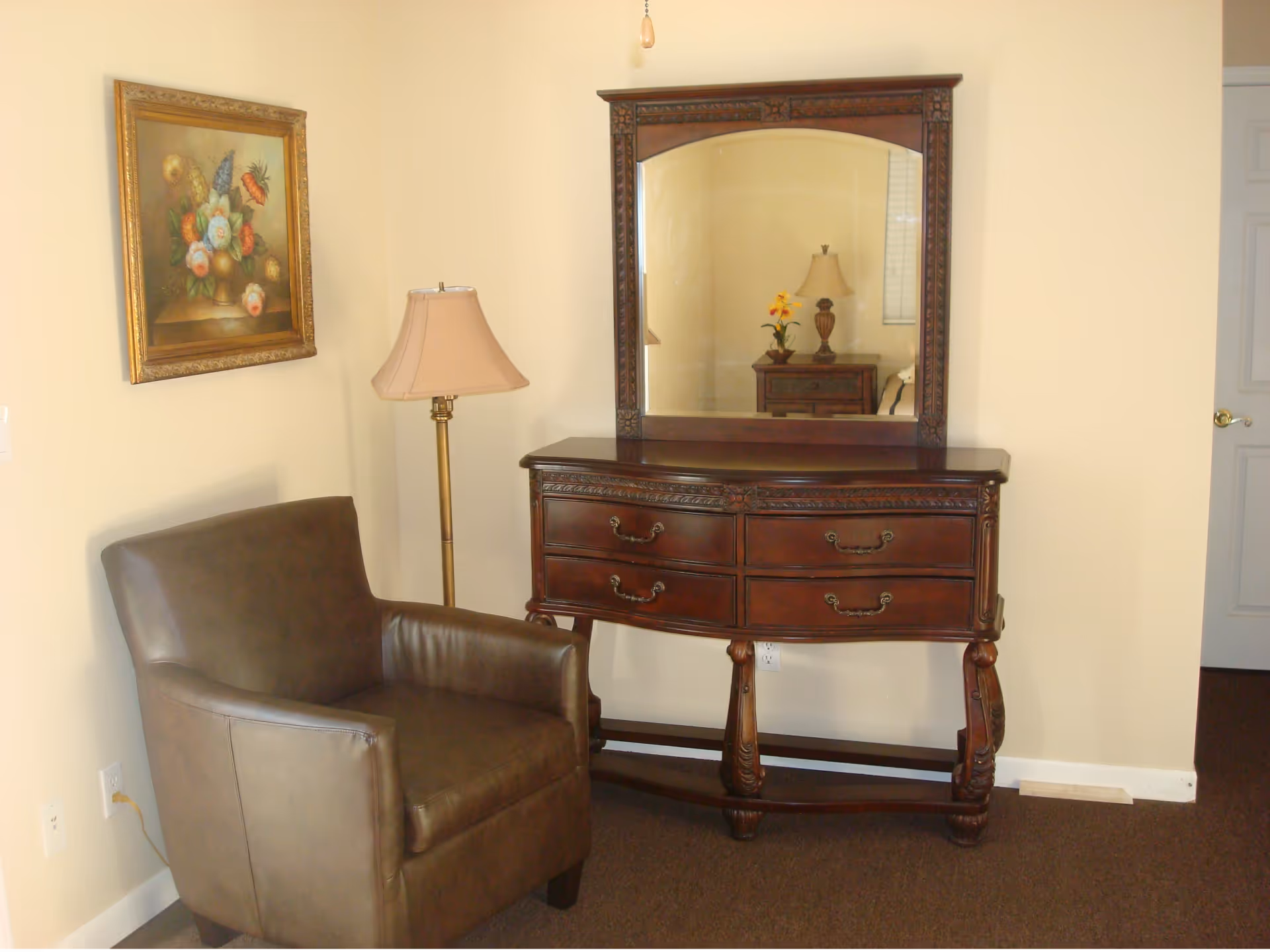 A cozy corner of a room featuring a brown leather armchair, a tall floor lamp with a beige shade, and a dark wooden dresser with an ornate mirror. A framed floral painting hangs on the wall above the armchair. The room has beige walls and brown carpet.