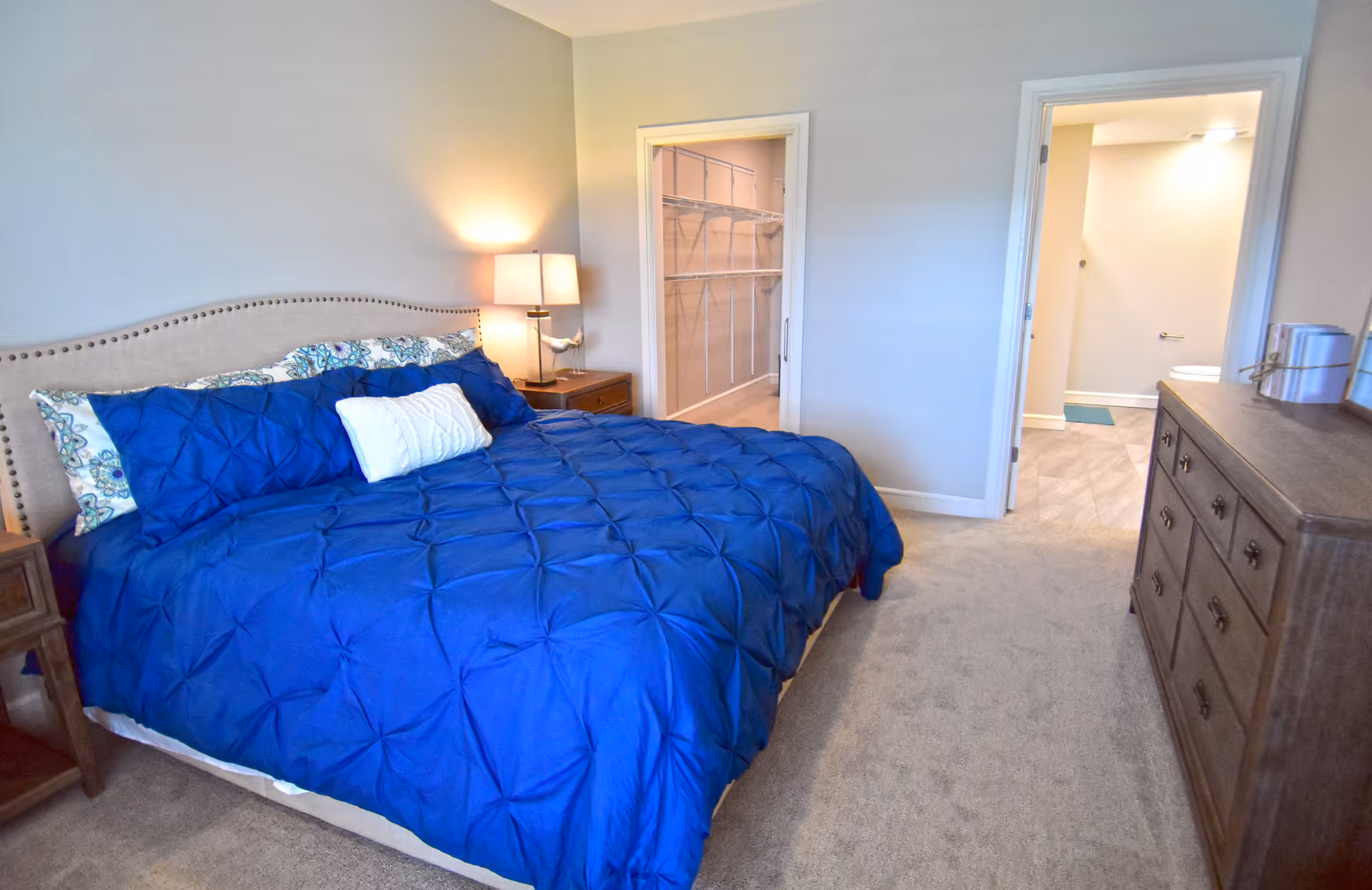A bedroom with a large bed covered in a blue comforter and decorative pillows. There is a wooden nightstand with a lamp and a bird figurine next to the bed. Two doorways are visible; one leads to a walk-in closet with shelving, and the other leads to a bathroom. A wooden dresser with books and decor is on the right side of the room.