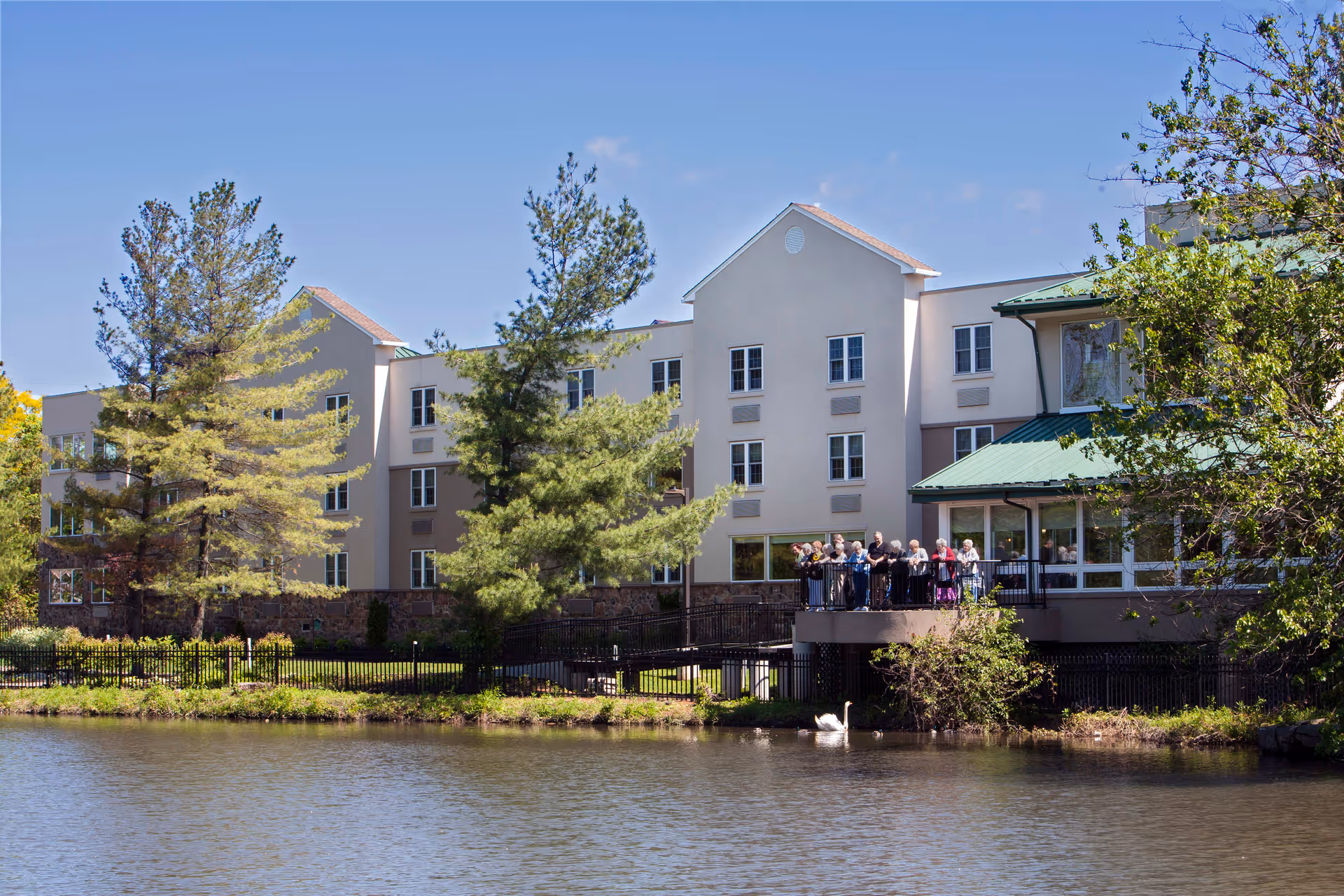 Exterior view of a senior living facility named Commonwealth Senior Living At Willow Grove, showing a multi-story building with beige walls and green roofs. There are several tall trees around the building and a body of water in the foreground with a swan swimming. A group of elderly people is gathered on a balcony overlooking the water.