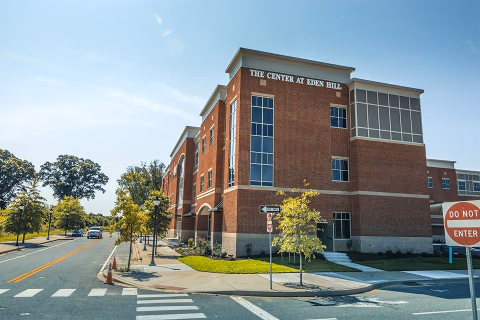 Exterior view of a large brick building named The Center At Eden Hill on a sunny day, with a street and sidewalk in the foreground, trees lining the street, and traffic signs including a 'Do Not Enter' and 'One Way' sign.