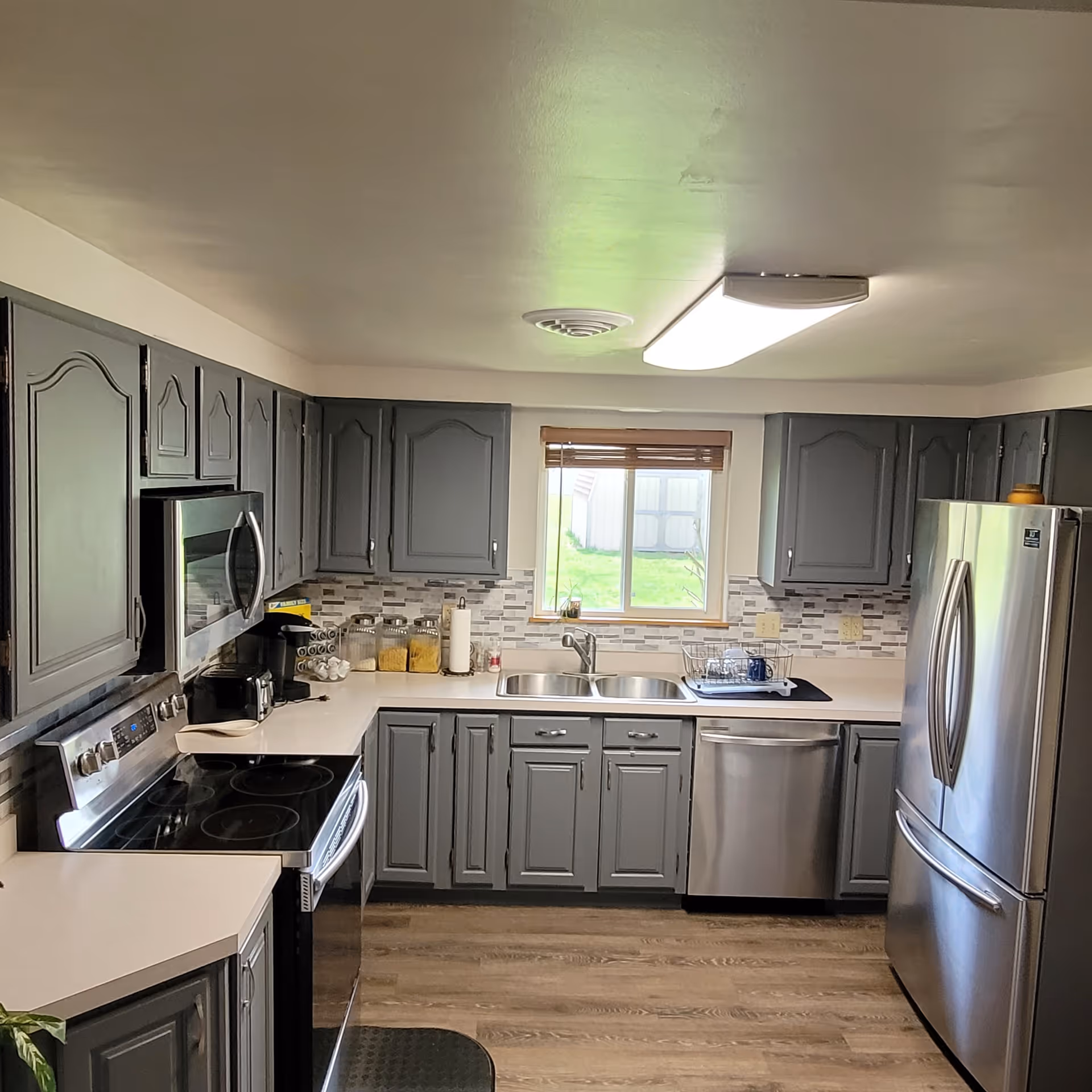 A modern kitchen with gray cabinets, stainless steel appliances including a refrigerator, microwave, stove, and dishwasher. There is a double sink under a window with a view of a green lawn outside. The countertops are light-colored with various kitchen items such as jars, a coffee maker, and a dish rack.
