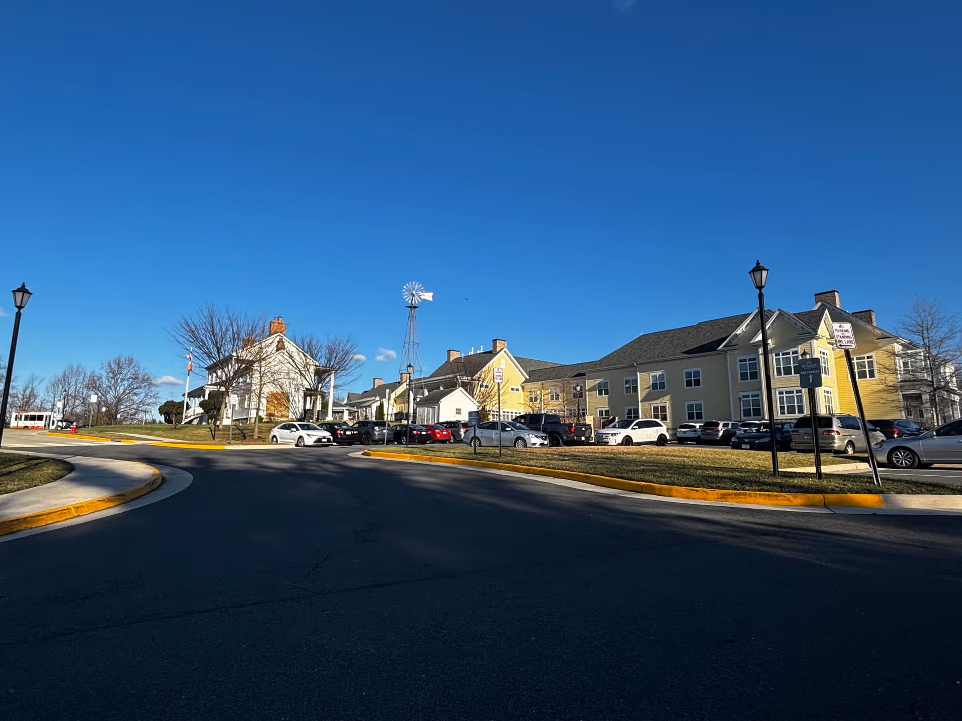Wide exterior view of Sunrise at Silas Burke House facility showing a large yellow and white building with multiple windows, a parking lot filled with cars, a windmill structure, street lamps, and a clear blue sky.