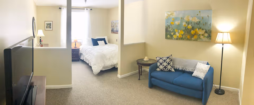 A cozy living space in an assisted living facility featuring a blue loveseat with decorative pillows and a throw blanket, a floor lamp, a round side table with a candle, and a floral painting on the wall. Adjacent to the living area is a bedroom with a neatly made bed, nightstand with a lamp, and a window with white curtains allowing natural light to brighten the room.