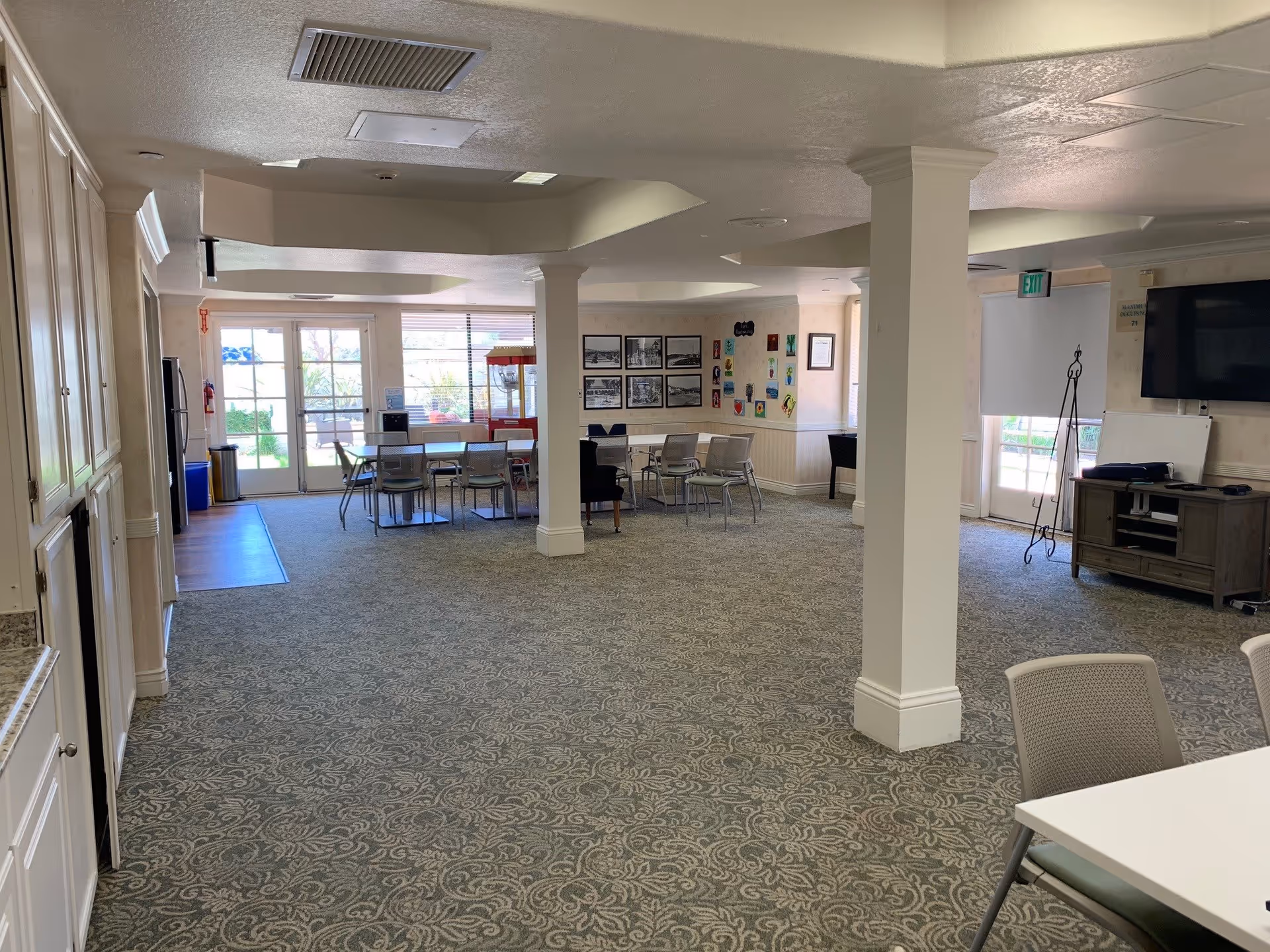A spacious common area in a senior living facility with patterned carpet, white pillars, and multiple tables and chairs arranged for group activities. There are large windows and glass doors letting in natural light, a popcorn machine near the back, framed pictures and colorful artwork on the walls, a TV mounted on the wall, and a cabinet with a whiteboard and other items.