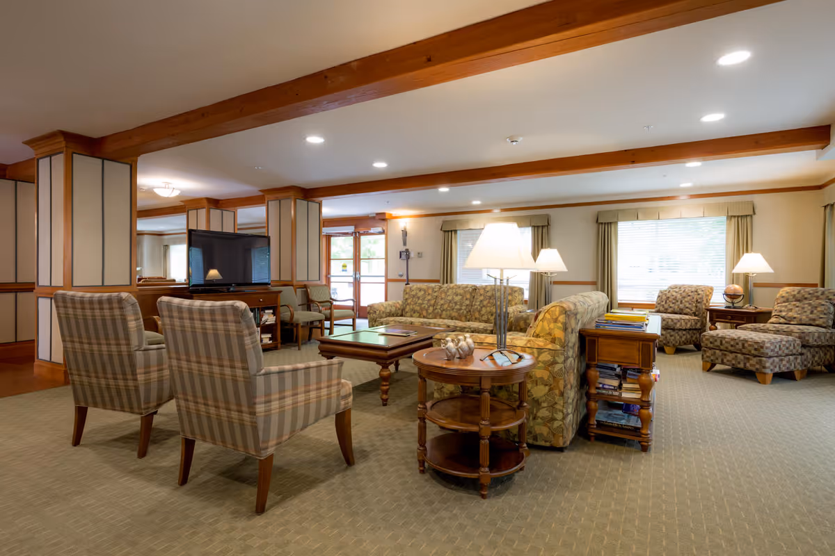 A spacious living room area with patterned armchairs and sofas arranged around wooden coffee tables. The room has beige carpeting, wooden beams on the ceiling, and several lamps providing warm lighting. Large windows with curtains allow natural light to enter, and a flat-screen TV is visible near the back of the room.