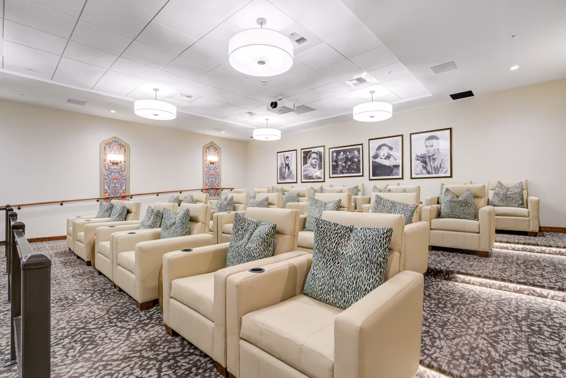 A small theater room with three rows of beige leather armchairs, each with a patterned cushion. The room has carpeted floors, soft lighting from ceiling fixtures, and framed black and white portraits on the far wall.