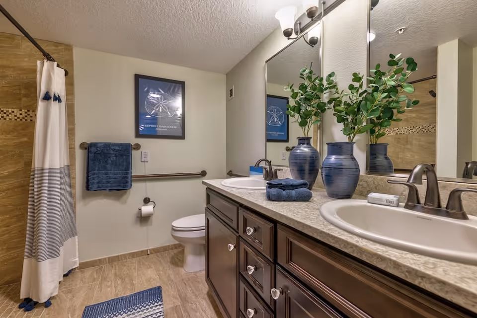 A bathroom with a double sink vanity featuring dark wood cabinets and a granite countertop. Two blue vases with green leafy plants and folded blue towels are placed on the countertop. A large mirror with two wall-mounted light fixtures is above the sinks. To the left, there is a toilet with a blue towel hanging on a grab bar and a framed picture on the wall. A shower with a white and blue striped curtain is visible in the background. The floor and shower walls are tiled in beige tones.