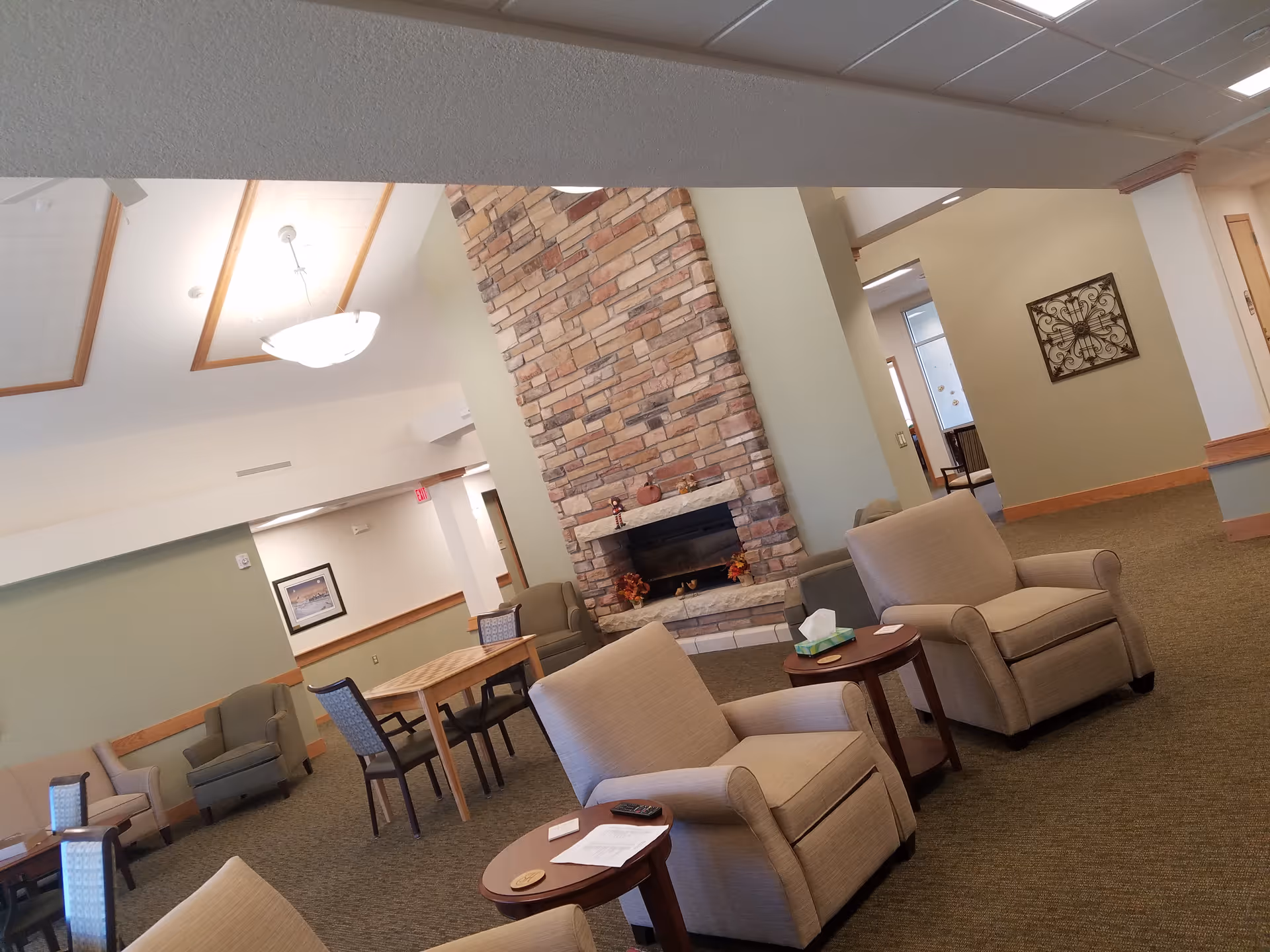A cozy living room area in Hillcrest Living Campus featuring a stone fireplace in the center, surrounded by beige armchairs and small wooden tables. The room has a carpeted floor, light-colored walls, and a ceiling with recessed lighting and a hanging light fixture. There are additional chairs and tables in the background, along with framed artwork on the walls.