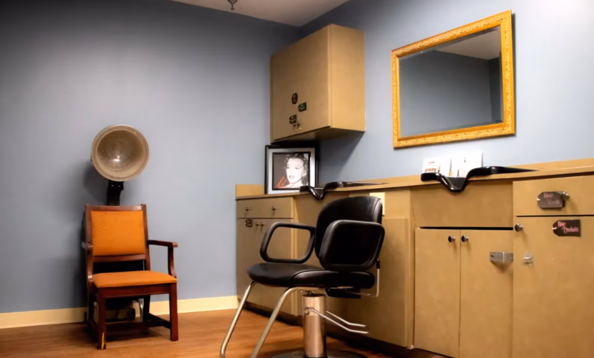 Interior of a hair salon area with a black salon chair, a vintage hair dryer chair with an orange cushion, beige cabinets with a sink, a large mirror with a gold frame, and a black and white framed photo on the counter.