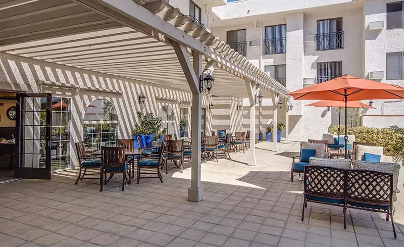 Outdoor patio area at Dania's Senior Home with tables and chairs under a pergola and an orange umbrella, adjacent to a multi-story building with windows and balconies.