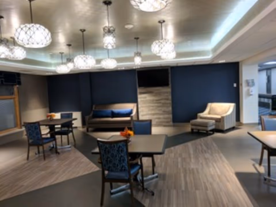 A modern common area with multiple tables and chairs, a sofa with blue cushions, an armchair with an ottoman, and several hanging pendant lights. The walls are painted dark blue and the floor has a mix of wood and tile patterns.
