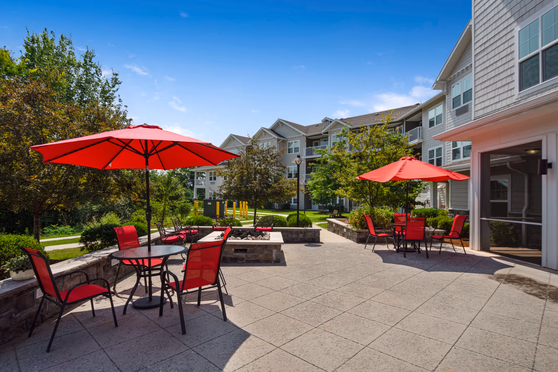 Sunlit outdoor patio with red umbrellas, tables, chairs and a fire pit in front of a multi-story residential building.