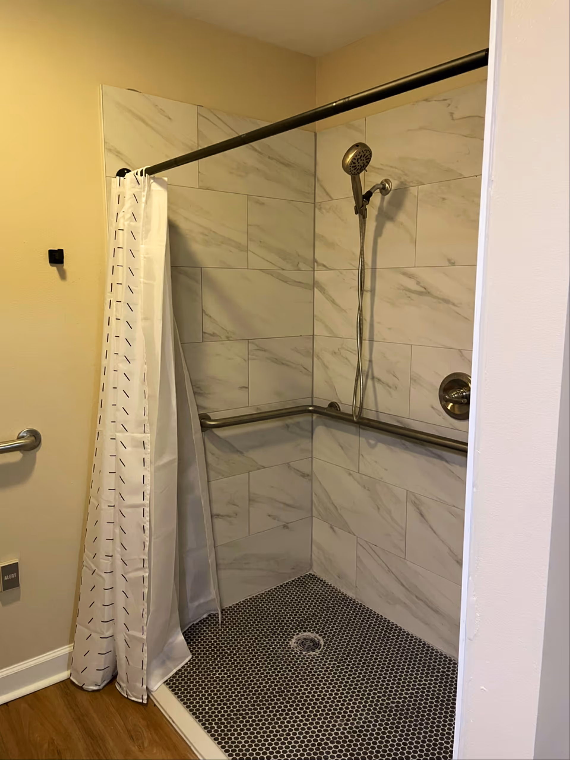 Tiled walk-in shower with a handheld showerhead, horizontal grab bars, and a patterned shower curtain in a bathroom.