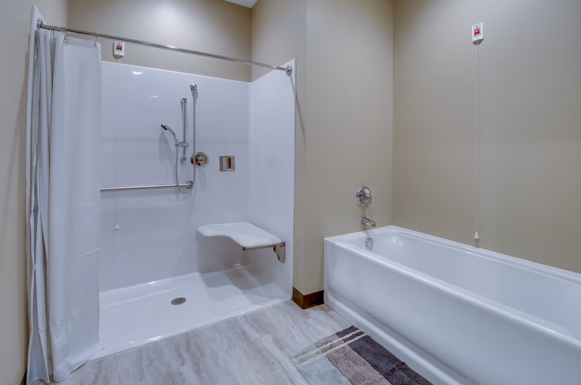 A clean and accessible bathroom featuring a walk-in shower with a fold-down seat, grab bars, and a handheld showerhead, alongside a standard bathtub. The walls are painted beige and the floor has light-colored tiles with a small rug near the bathtub.