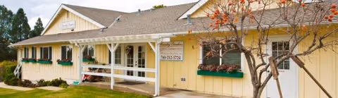 Yellow single-story senior living facility with white trim, planters, and a tree at the entrance.