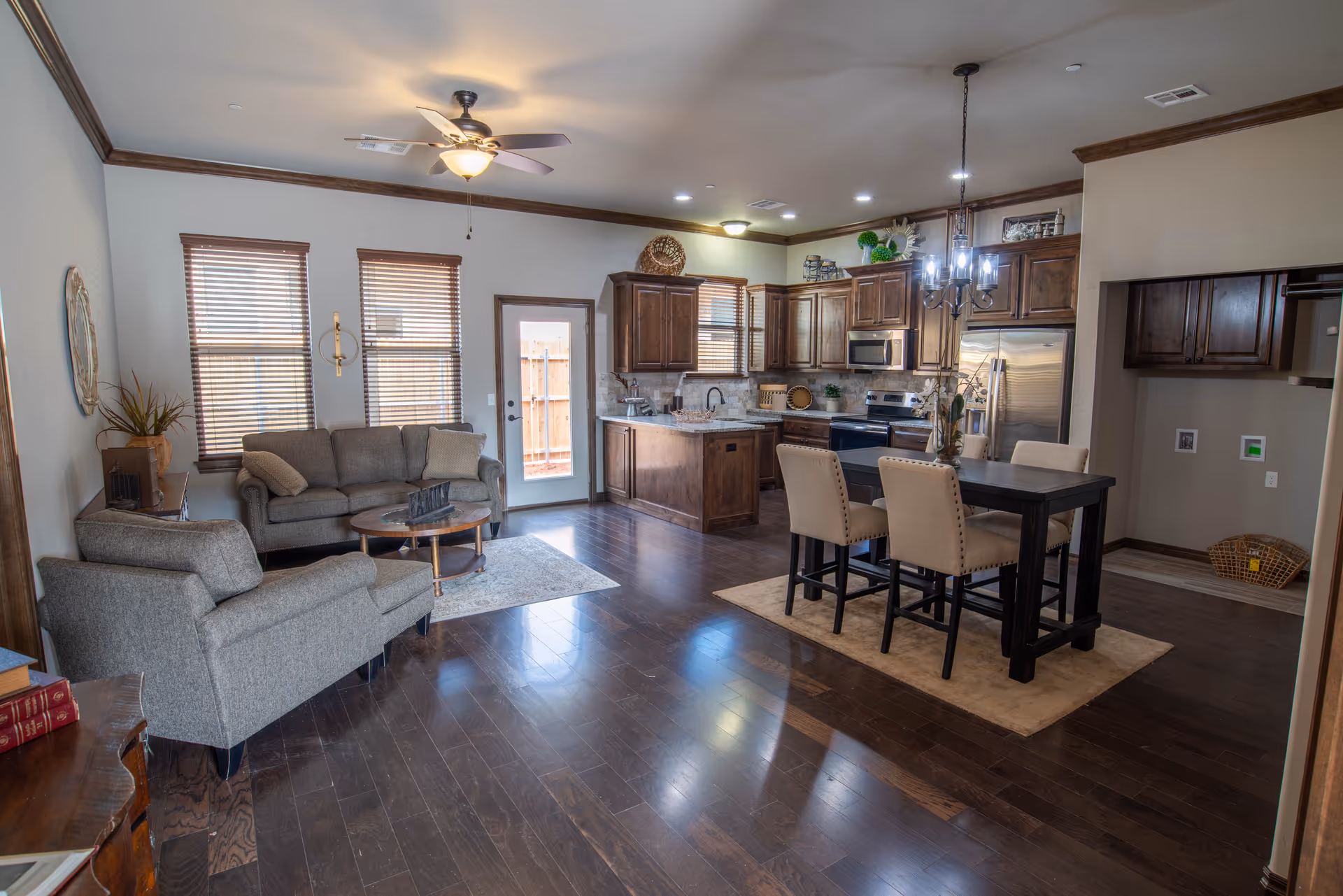 Open-concept living area featuring a seating area with a gray sofa and armchair, a round wooden coffee table on a small rug, and a kitchen with wooden cabinets, stainless steel appliances, and a dining table with four upholstered chairs under a chandelier. The room has dark hardwood floors, three windows with blinds, and a door leading outside.