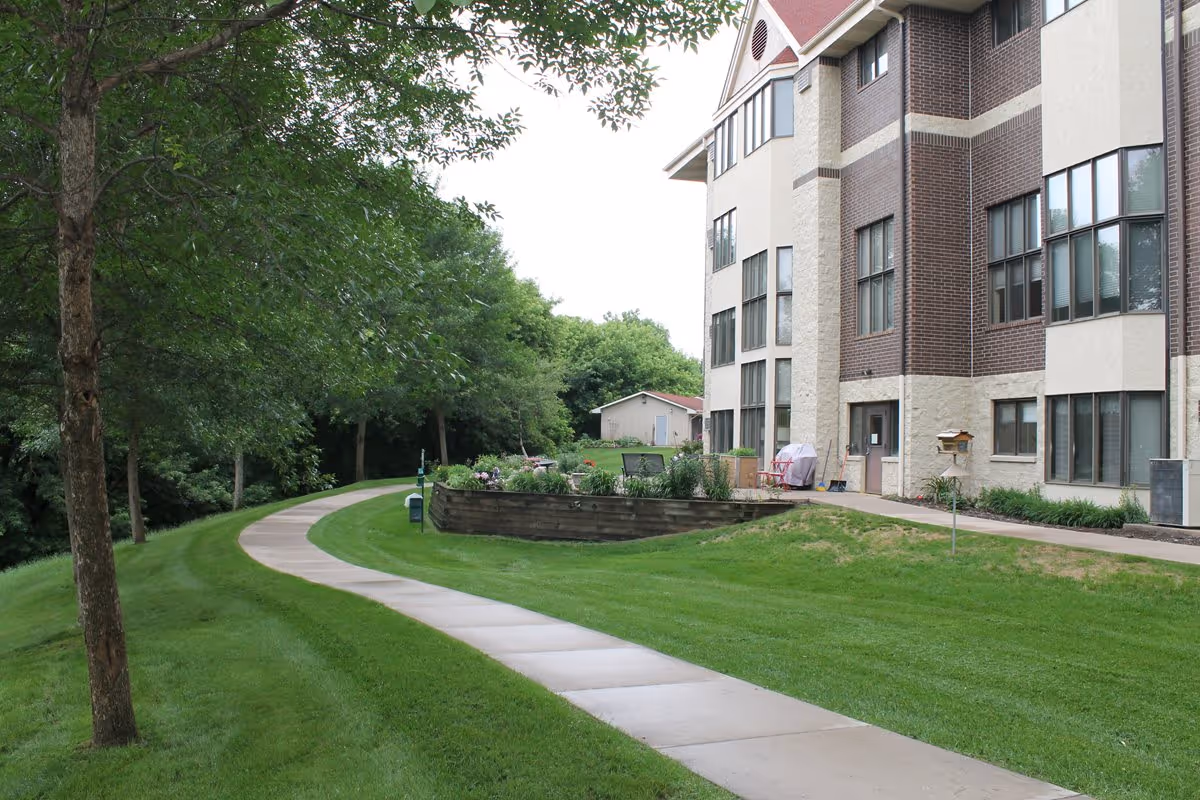 A paved walkway curves through a well-maintained grassy area with trees on the left and a multi-story building with large windows on the right. There is a small garden bed with flowers and patio furniture near the building, and a small shed is visible in the background.