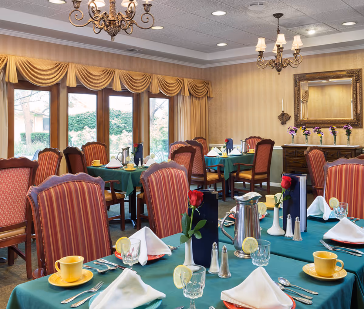 A dining room with tables covered in green tablecloths, set with yellow cups and saucers, glassware with lemon slices, silverware, white folded napkins, and small vases each holding a single red rose. The room has striped upholstered chairs, large windows with gold drapes, chandeliers, and a sideboard with a mirror and flower vases.
