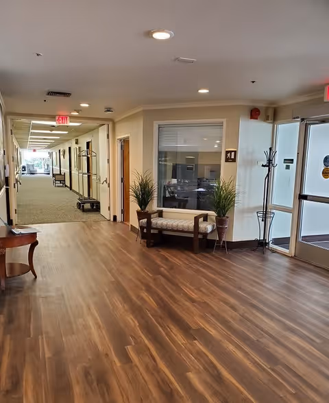 Interior view of a senior living facility hallway with wood flooring and beige walls. There is a cushioned bench with two potted plants on either side against the wall, a coat rack near the glass exit door, and a long carpeted corridor with handrails and some furniture visible in the distance.