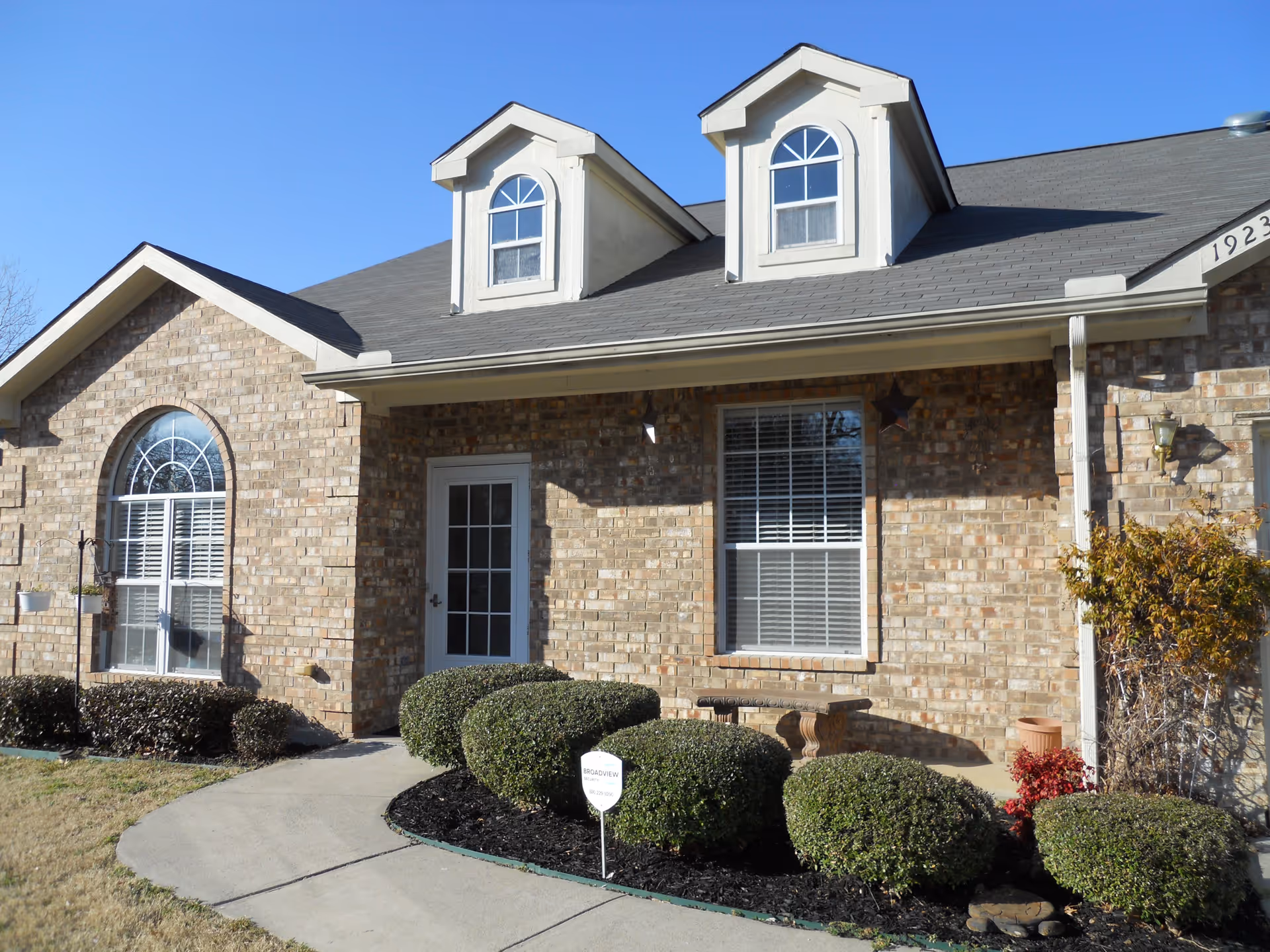 Exterior view of a brick residential building with two dormer windows on the roof, a white door, and two large windows. There is a curved concrete walkway leading to the door, surrounded by neatly trimmed bushes and a small garden area. The sky is clear and blue.