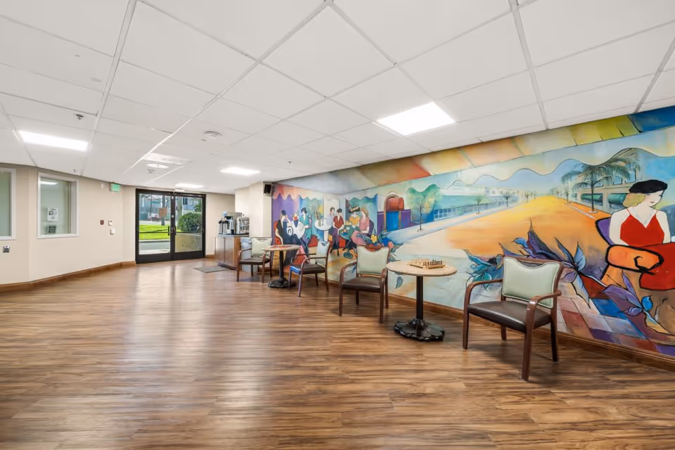 Spacious interior common area with wood floors, chairs and small tables along a colorful wall mural and glass entrance doors.