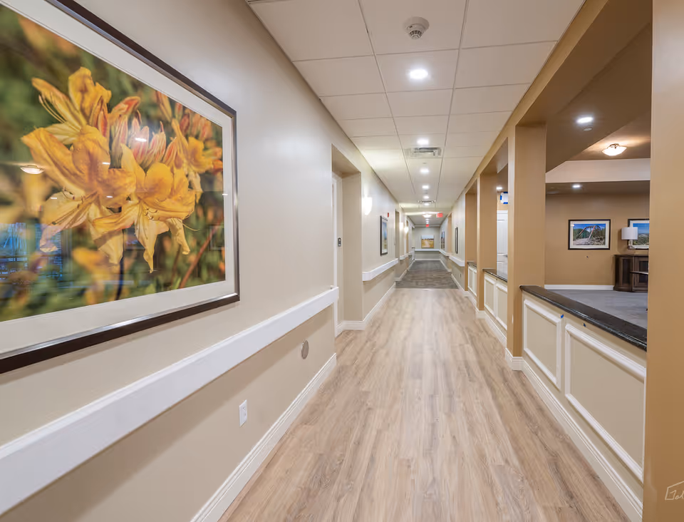 A long, well-lit hallway with light wood flooring and beige walls. The hallway has white handrails on both sides and framed floral artwork hanging on the left wall. On the right side, there is an open area with beige columns and a countertop, leading to a room with additional framed pictures and a lamp.