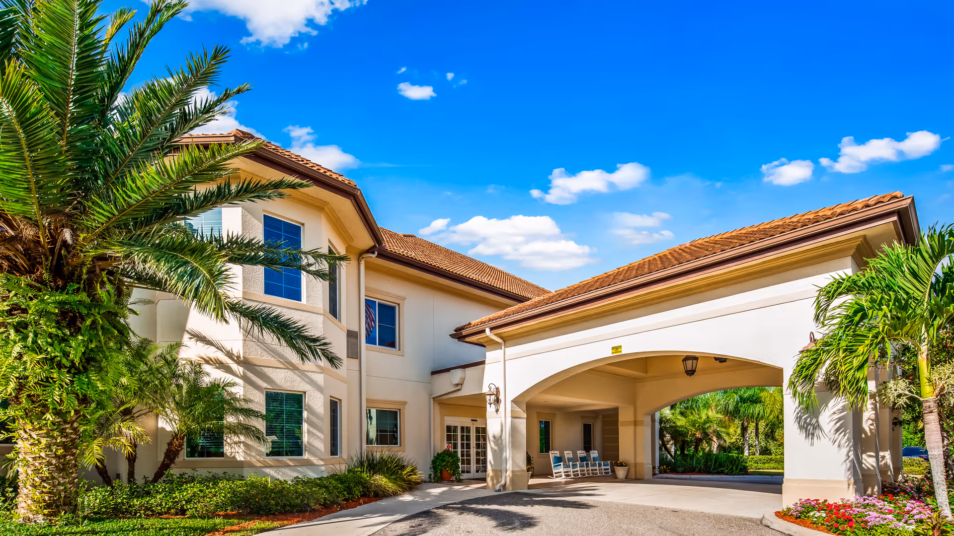 Exterior view of The Windsor of Venice senior living facility showing a two-story building with beige walls and a red-tiled roof under a bright blue sky with scattered clouds. There are palm trees and well-maintained landscaping around the entrance, which features a covered driveway with rocking chairs.