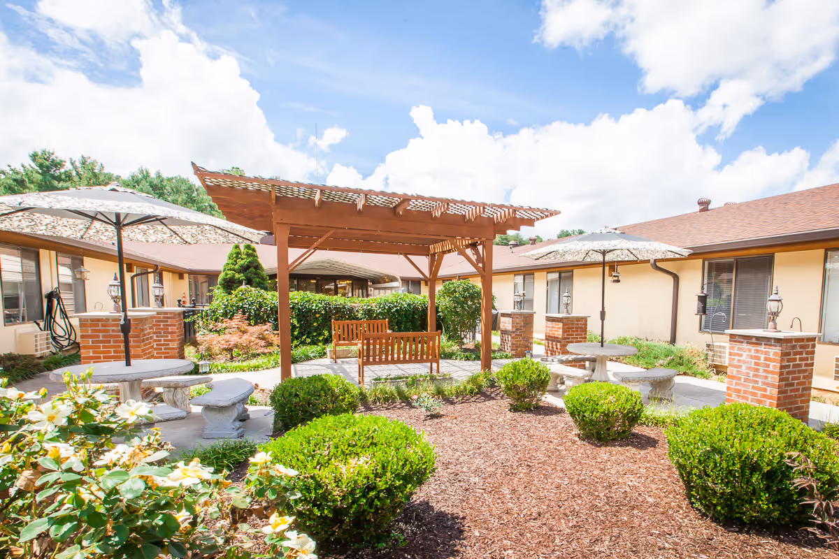 Outdoor courtyard area with a wooden pergola, benches, round stone tables with umbrellas, surrounded by bushes and flowers, and single-story building wings in the background under a partly cloudy sky.