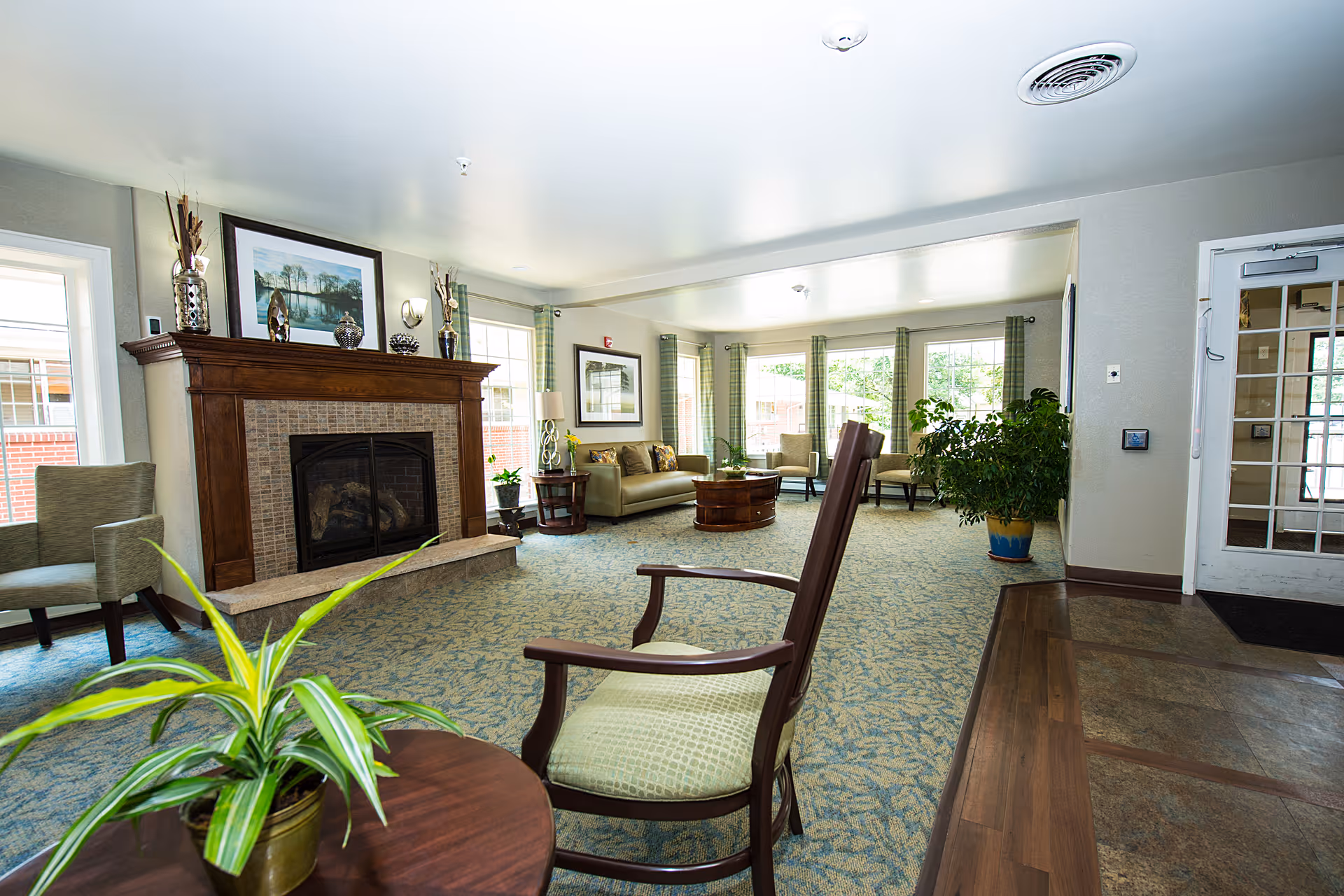 A bright and spacious living room area with a fireplace featuring a wooden mantle decorated with vases and a framed picture above it. The room has multiple seating options including armchairs and sofas arranged around a central wooden coffee table. Large windows with green curtains allow natural light to fill the space, and there are several potted plants adding greenery to the room. The floor is carpeted with a patterned design, and there is a door with glass panels on the right side.