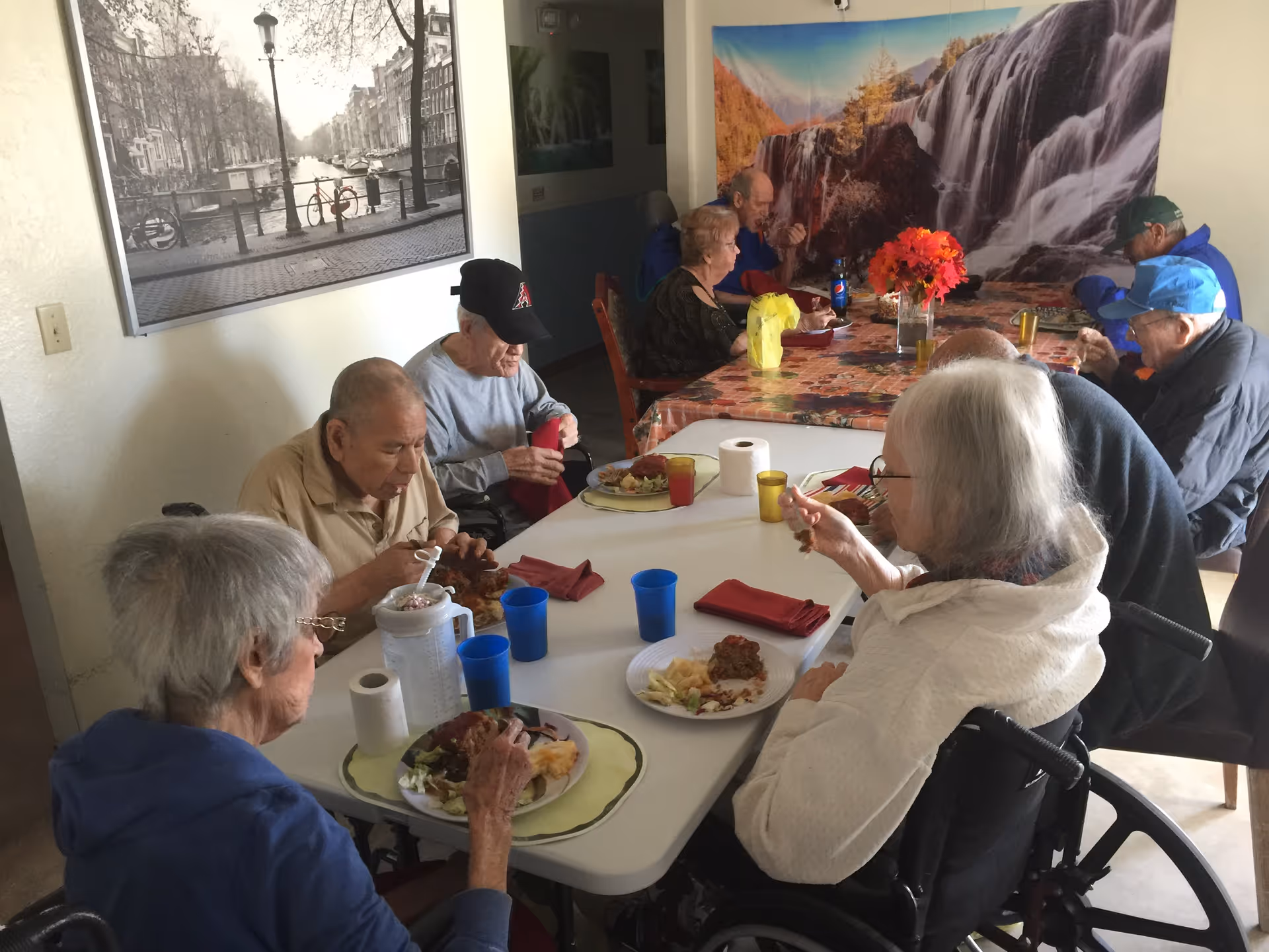 A group of elderly people sitting around two tables in a dining area, eating a meal together. The room has wall art including a black and white photo of a canal and a large colorful waterfall mural. The tables have plates of food, cups, napkins, and a vase with red flowers.