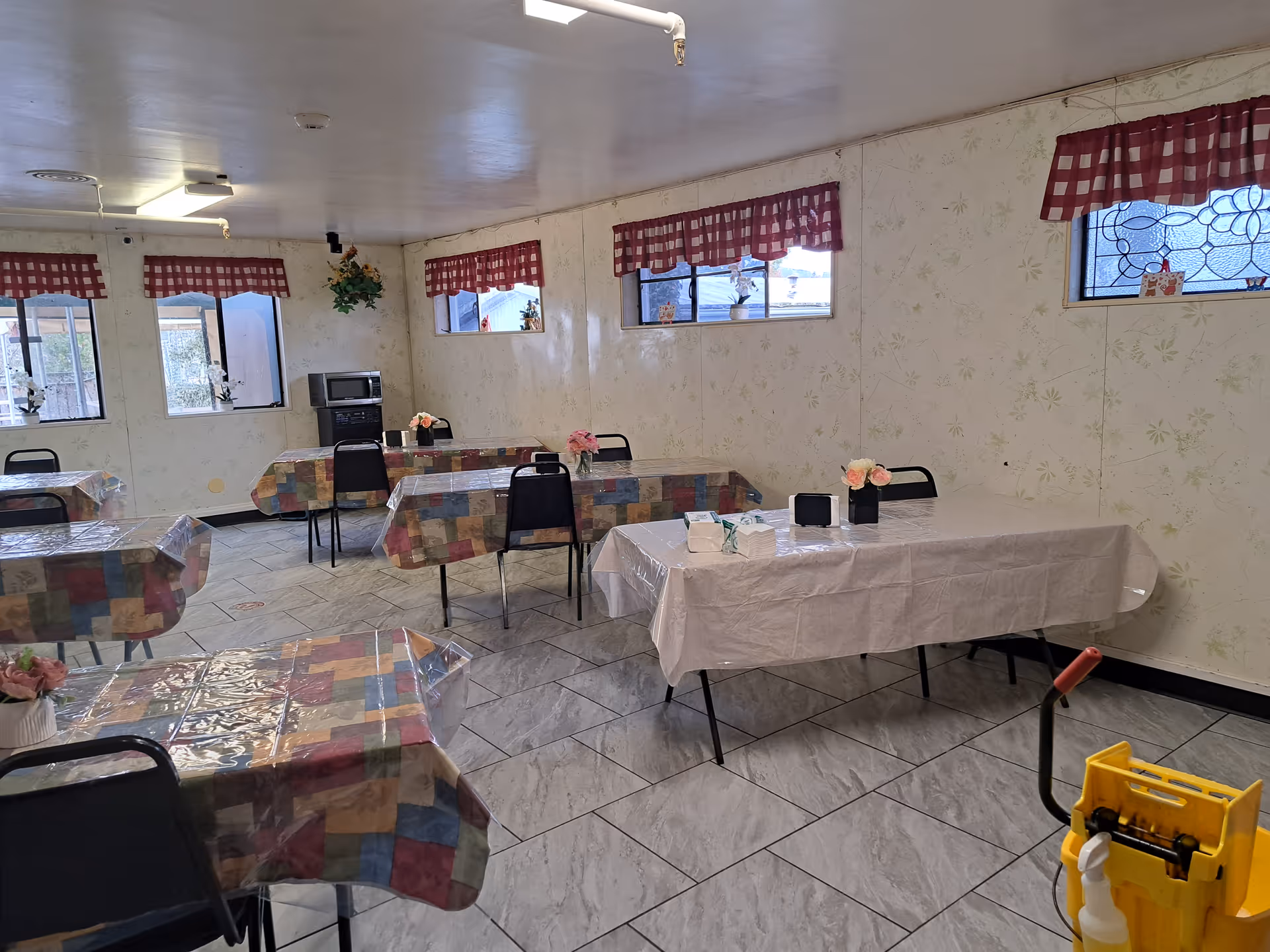 Communal dining room with several tables covered in colorful patterned tablecloths and plastic covers, chairs, small flower centerpieces, and a mop bucket in the foreground.