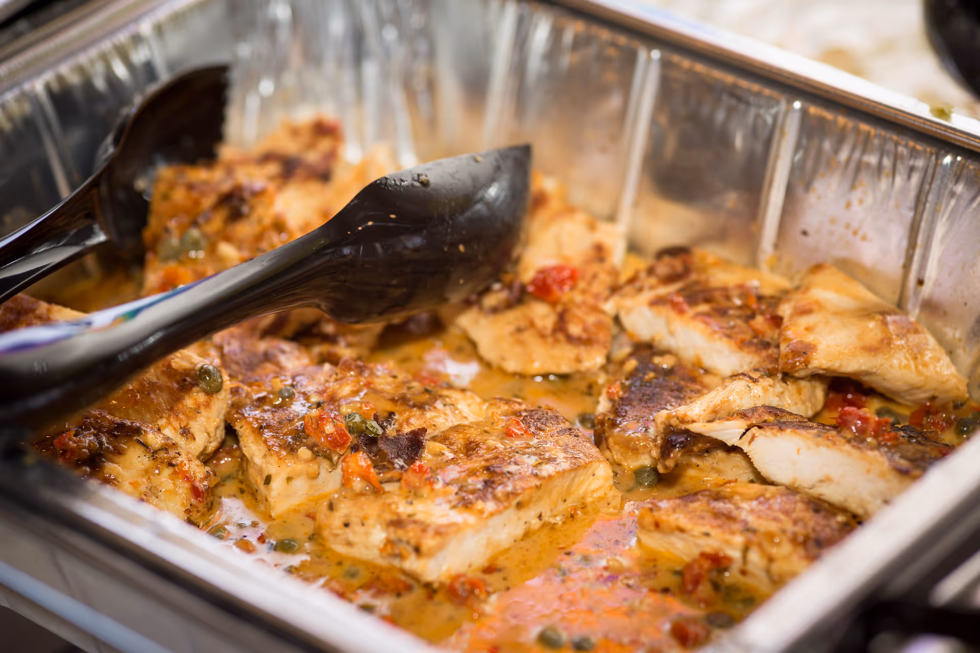 A metal buffet tray of cooked, sauced chicken pieces with black serving tongs.