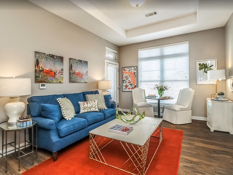 Bright living room with a blue sofa, red rug, coffee table, and two white chairs by a large window.