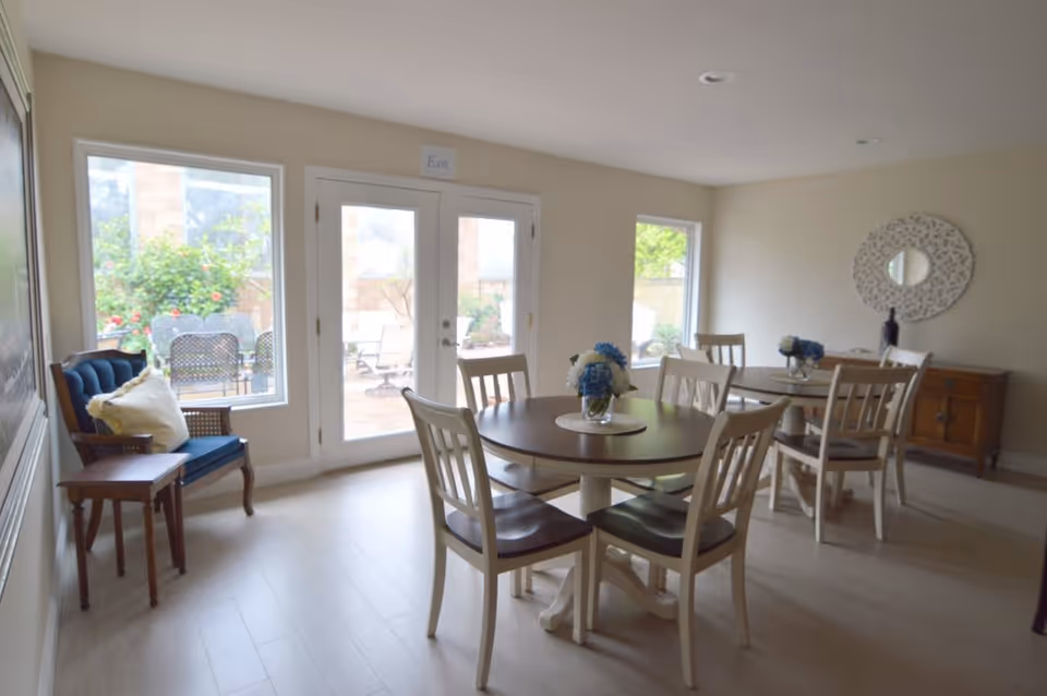 A bright dining room with two round wooden tables, each surrounded by four chairs. Each table has a small floral centerpiece. There is a blue cushioned armchair with a pillow near a window and a small wooden side table next to it. The room has light-colored walls and flooring, with large windows and glass doors leading to an outdoor patio area with garden plants visible outside. A decorative round mirror hangs on the wall above a wooden sideboard.