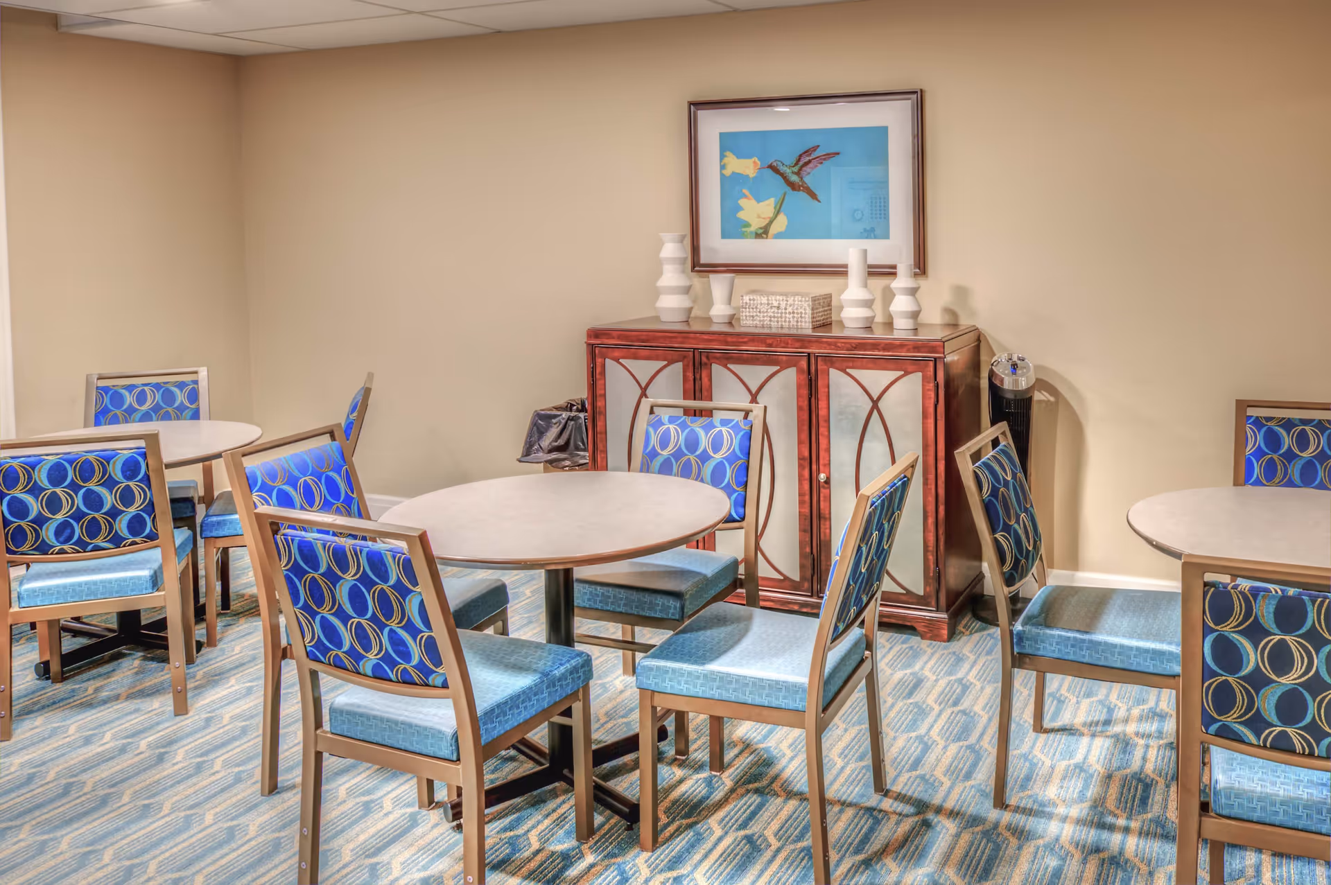 A small dining area with round tables and chairs featuring blue patterned upholstery. Against the beige wall is a wooden cabinet with decorative vases and a framed picture of a hummingbird and flowers above it. The floor is carpeted with a blue and beige geometric pattern.