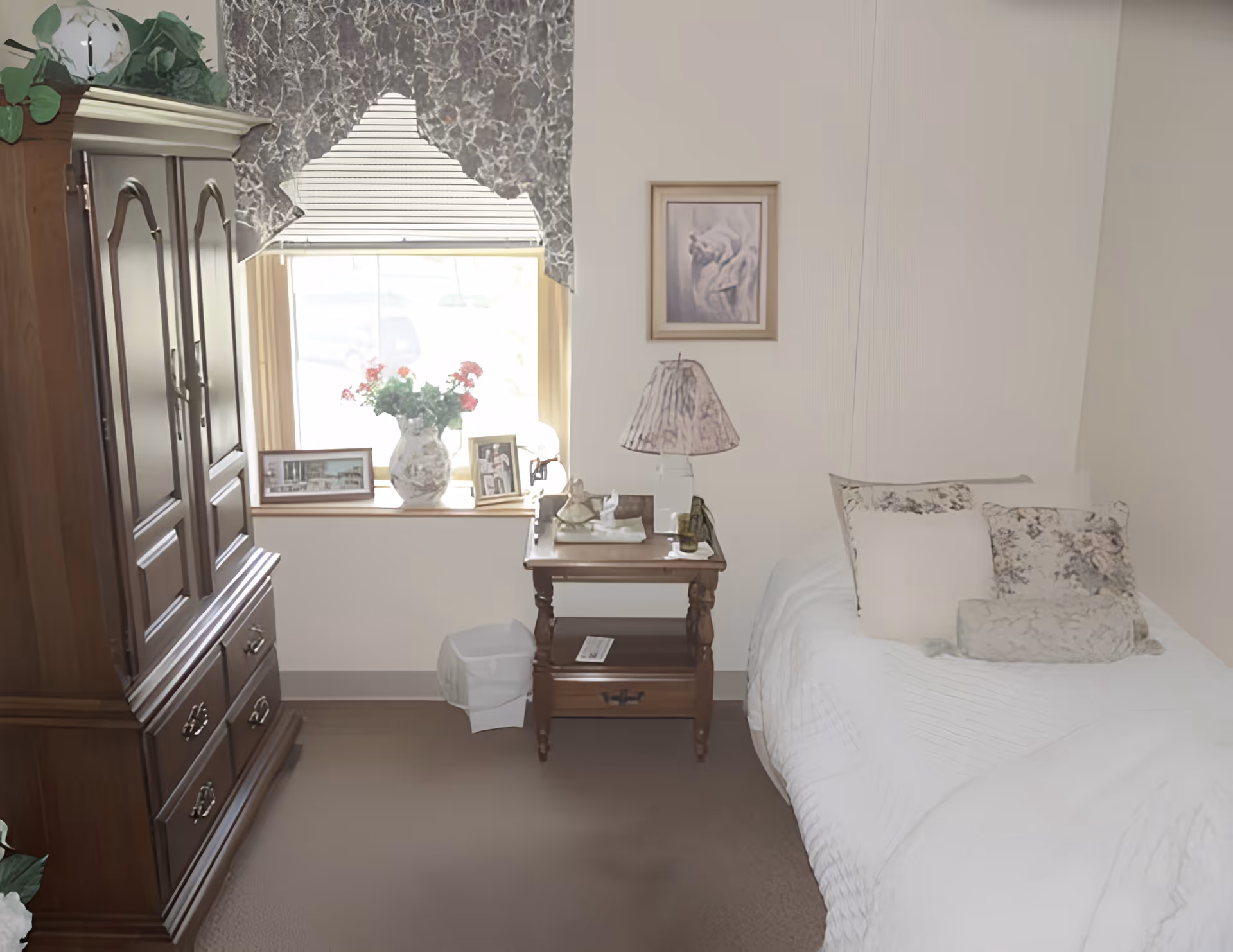 A cozy bedroom with a single bed covered in white bedding and decorative pillows. Next to the bed is a wooden nightstand with a lamp, a small decorative item, and a framed picture on the wall above. A window with a patterned valance lets in natural light, and a wooden dresser with drawers is positioned to the left. There is a vase with flowers and framed photos on the windowsill.
