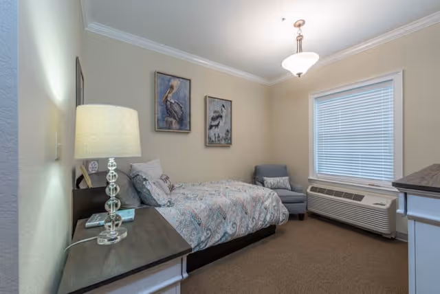 A cozy bedroom in an assisted living facility featuring a single bed with patterned bedding, a bedside table with a lamp and books, two framed bird paintings on the wall, a comfortable armchair with a pillow, a window with blinds, and a heating/cooling unit beneath the window.