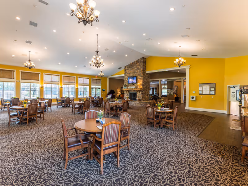 Spacious dining room with multiple round wooden tables and chairs arranged on a patterned carpet. The room features large windows with blinds, yellow walls, and several chandeliers hanging from a high ceiling. A stone fireplace with a mounted TV is visible in the center background, and a piano is placed near the fireplace.