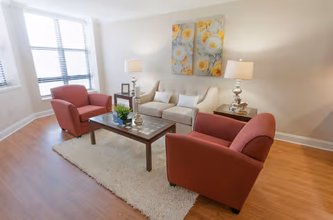 A bright living room with two red armchairs and a beige sofa arranged around a wooden coffee table on a light-colored rug. Two table lamps with white shades sit on side tables next to the sofa, and a three-panel floral painting hangs on the wall above the sofa. Large windows with blinds allow natural light into the room.