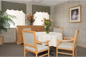 A small dining area with a round table covered by a white tablecloth surrounded by four wooden chairs with light-colored cushions. There is a wooden sideboard against the wall with a vase of flowers on top. Two windows with green valances and white blinds are behind the sideboard. A potted plant is in the corner, and a framed portrait hangs on the wallpapered wall.