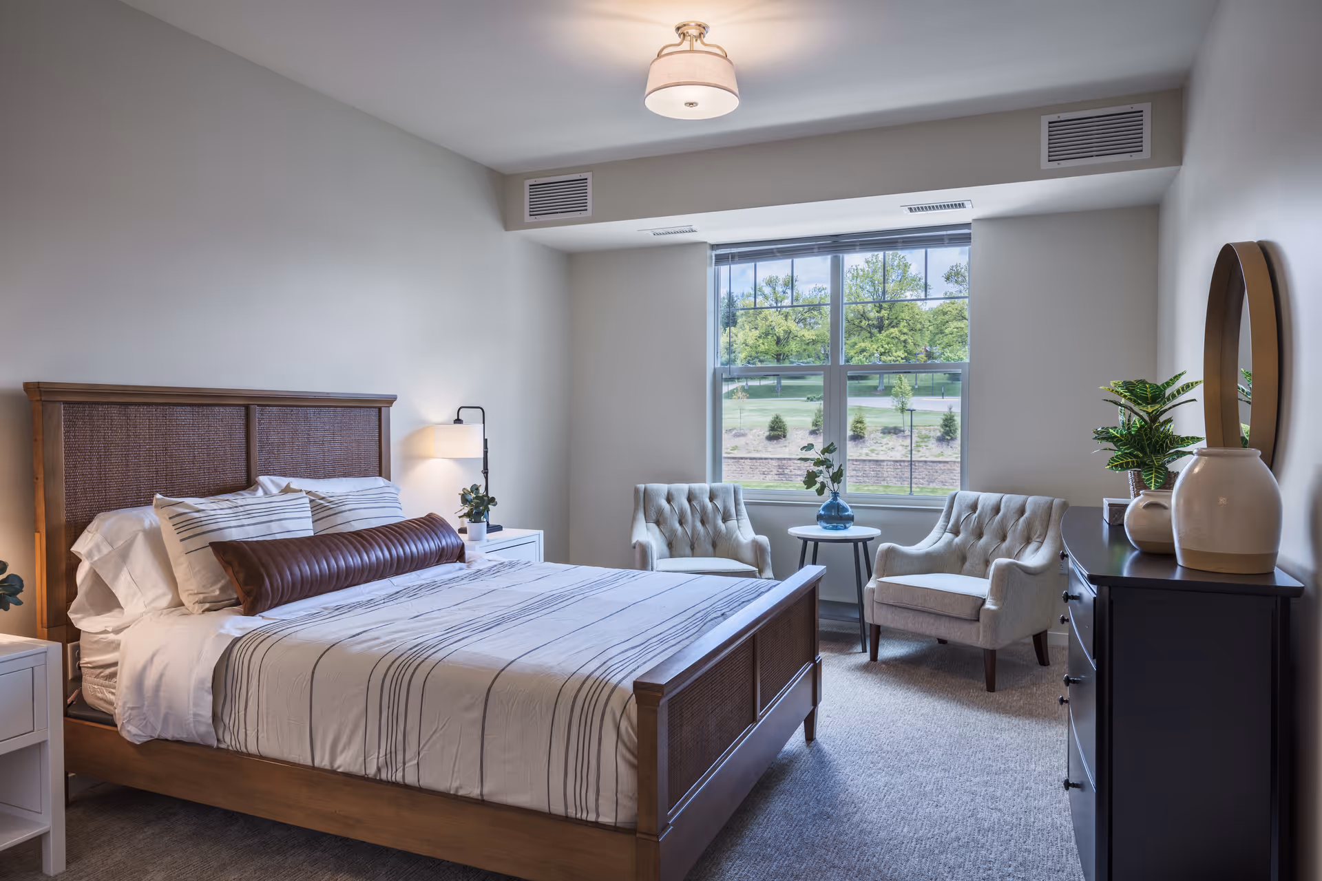 A bright and neatly arranged bedroom with a wooden bed frame, striped bedding, and a long cylindrical pillow. There are two white nightstands with lamps and plants on either side of the bed. Near a large window with a view of greenery outside, there are two cushioned armchairs with a small round table between them holding a blue vase with flowers. A dark dresser with a round mirror and decorative plants is on the right side of the room.